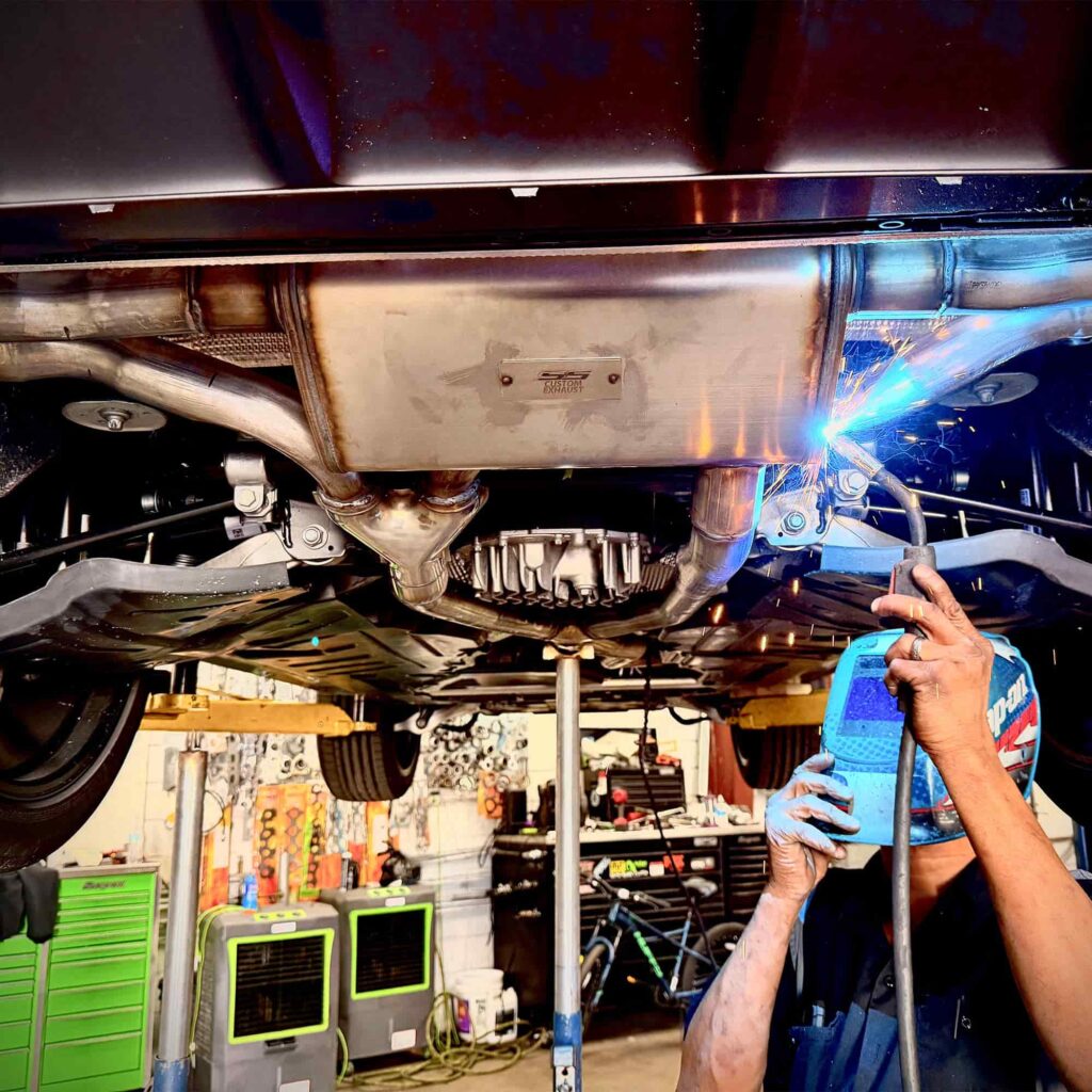Technician welding a custom exhaust system under a vehicle in an auto repair shop